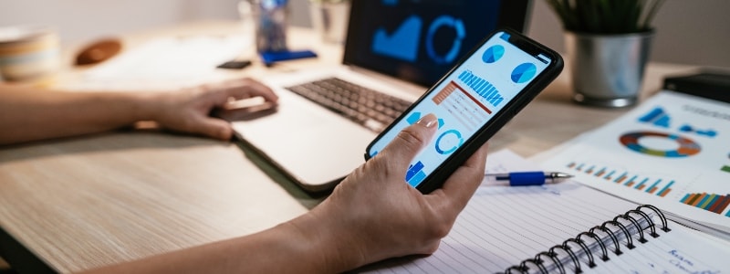 Close up of a hand holding a smartphone, with another hand in the background on a laptop, learning about fake tax preparers.