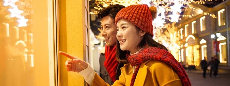 A young couple staring in the window of a store surrounded by Christmas lights and décor.