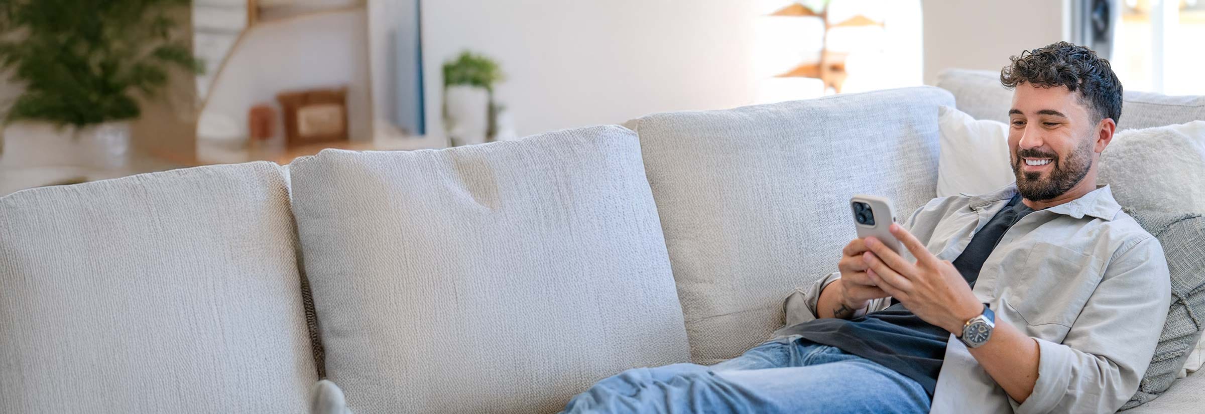 A man lying on the couch while using Hinge. His charming smile belies his sordid intentions to scam his match.