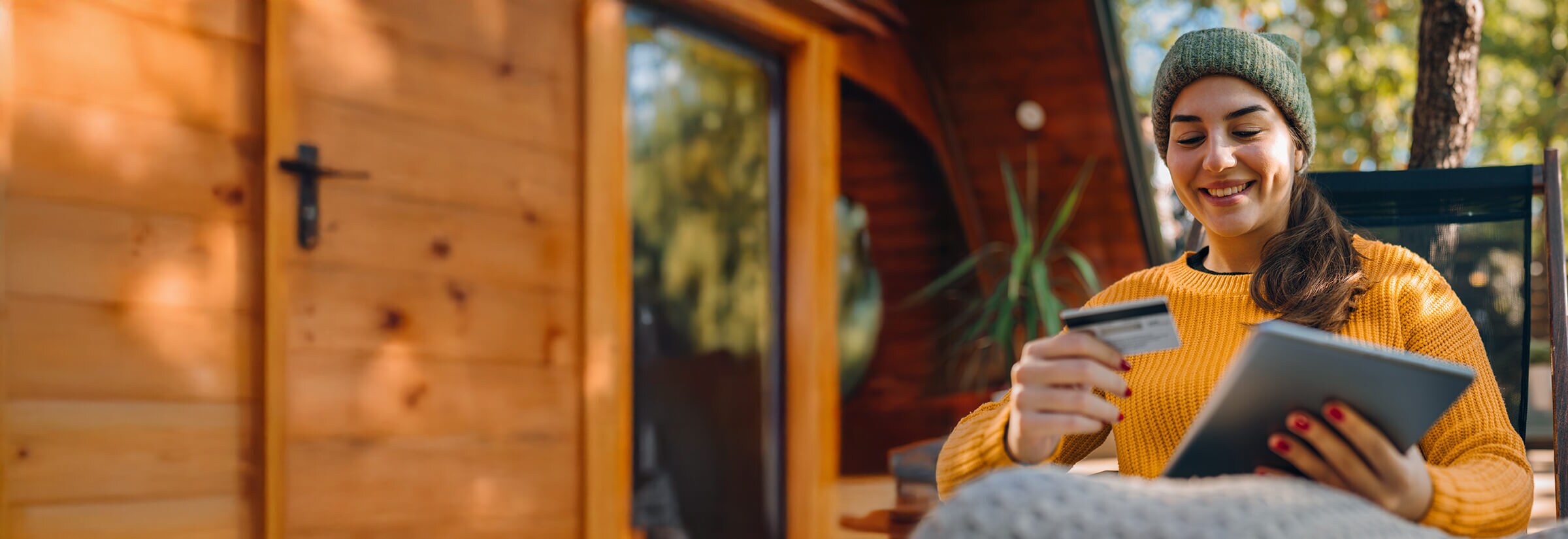 A woman sitting outside in a winter hat holds her credit card and tablet.