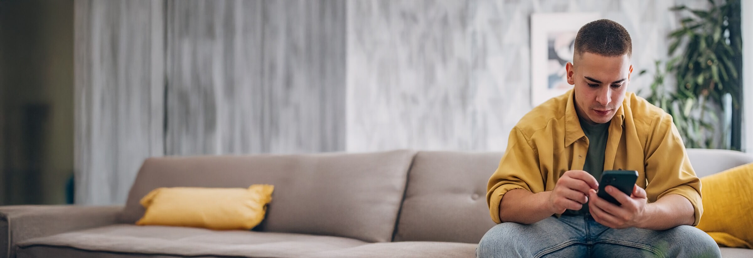 A man sits on a taupe sofa checking his phone for signs of hacking.
