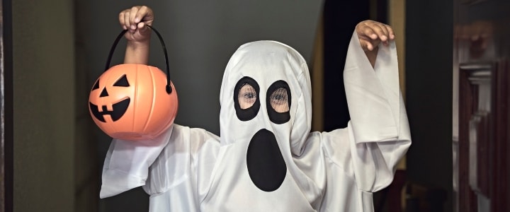 Little boy dressed in ghost costume holding a pumpkin-shaped Halloween candy basket.