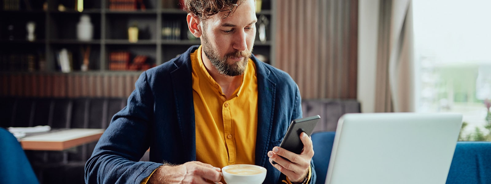 A man holding a cup of coffee looks at his phone for signs of identity theft to avoid.