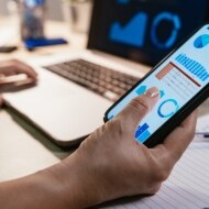 Close up of a hand holding a smartphone, with another hand in the background on a laptop, learning about fake tax preparers.