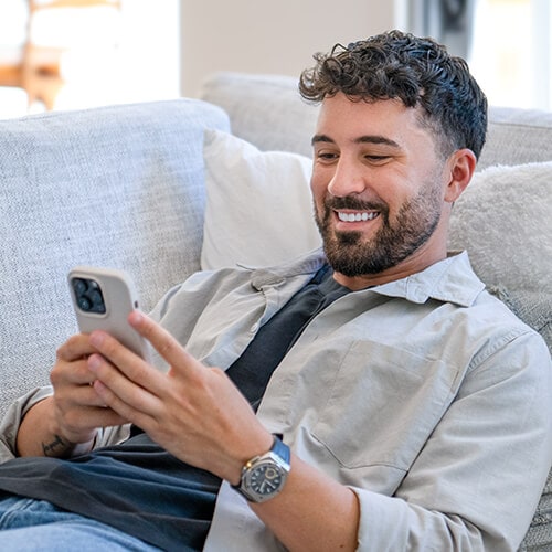 A man lying on the couch while using Hinge. His charming smile belies his sordid intentions to scam his match.