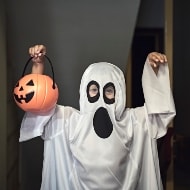 Little boy dressed in ghost costume holding a pumpkin-shaped Halloween candy basket.